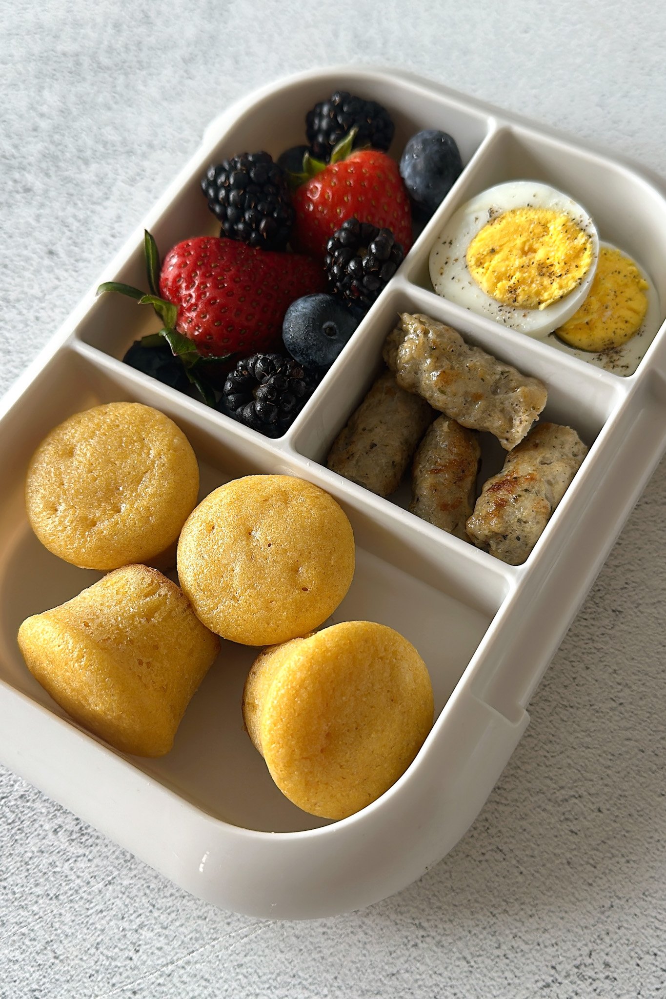 Mini corn muffins served inside a lunchbox with berries, boiled eggs, and sausages.