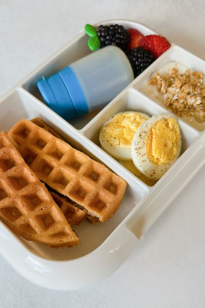 Eggless waffles, eggs, yogurt with granola, raspberries, raspberries, and maple syrup.