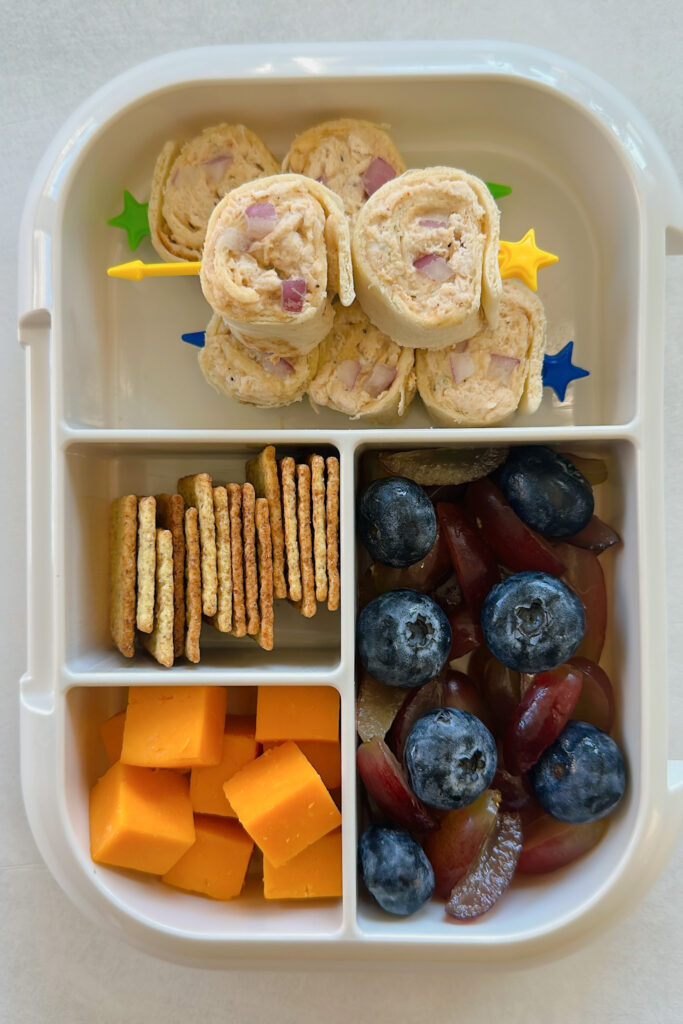 Chicken salad rollups served with crackers, cheese, blueberries, and grapes.