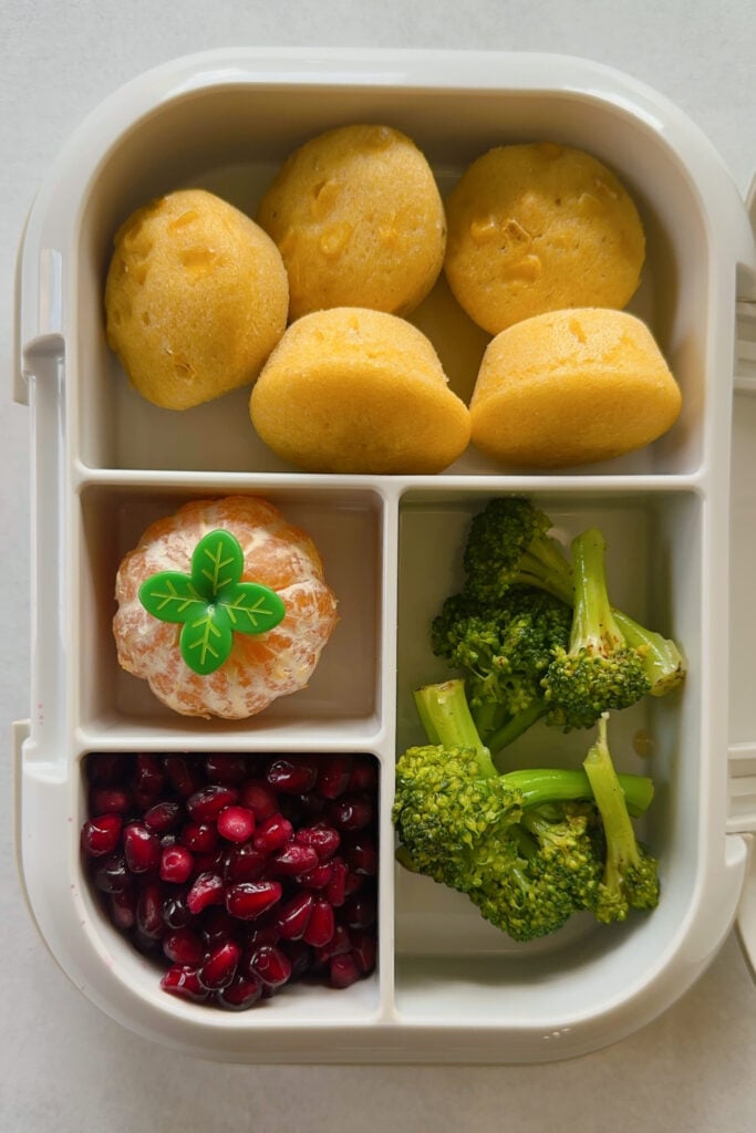 Corn muffins served with broccoli, mandarin, pomegranate seeds, and a that's it bar.