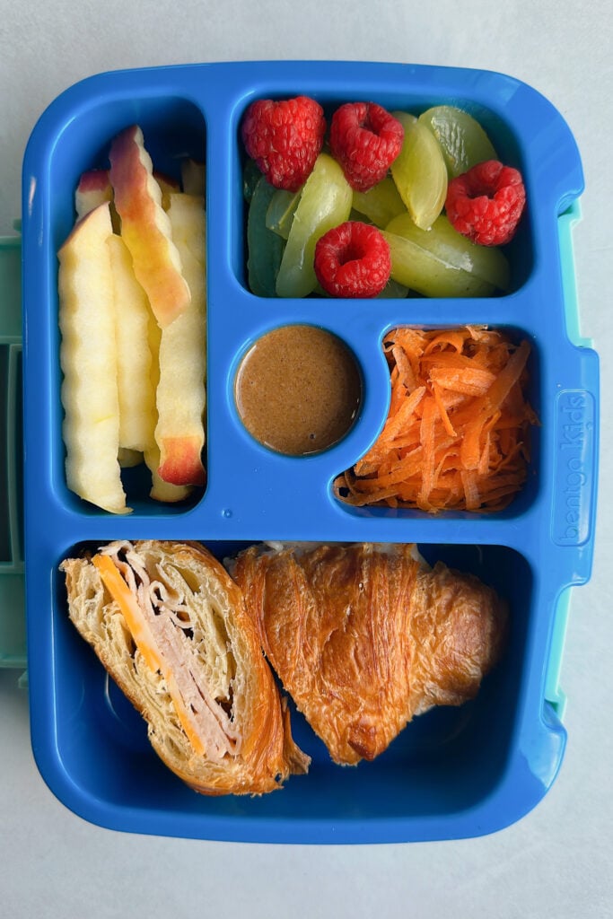 Turkey and cheese croissant served with apple's granola butter, grated carrots, grapes, and raspberries.