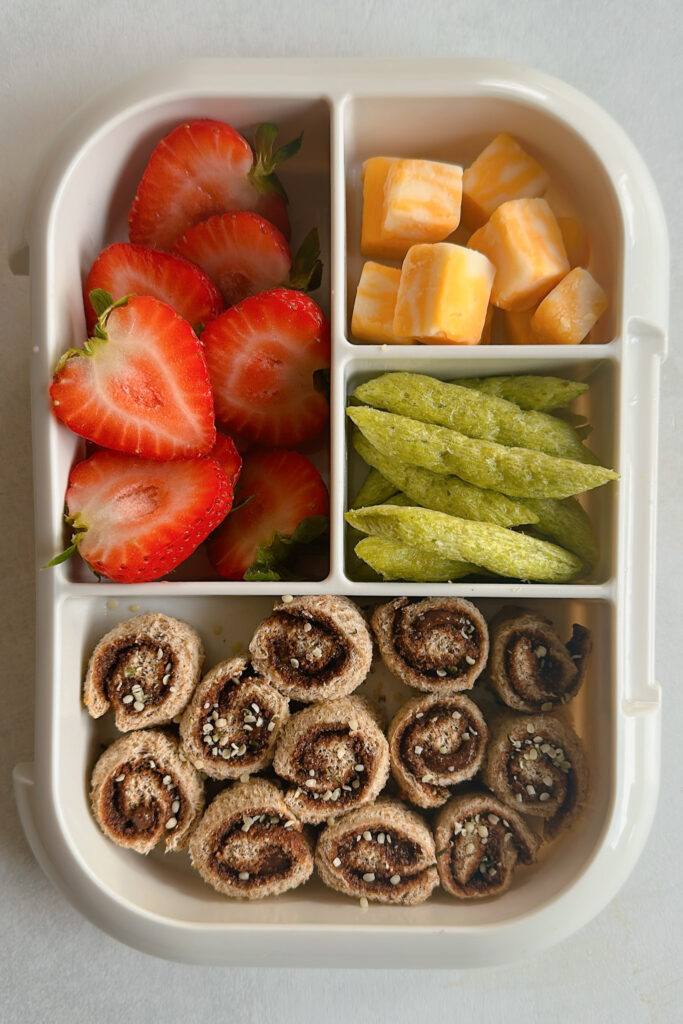 Granola butter rollups served with strawberries, cheese cubes, and snap peas crisps.