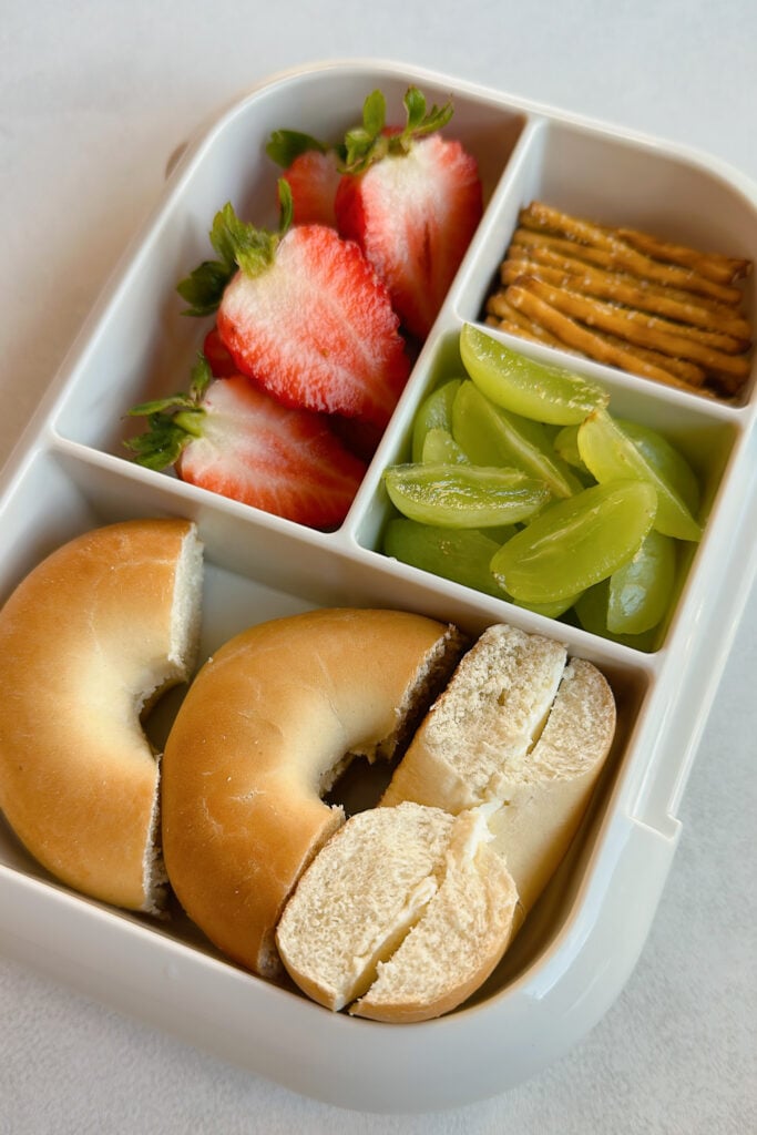 Mini bagel with cream cheese served with grapes, strawberries, and pretzels.