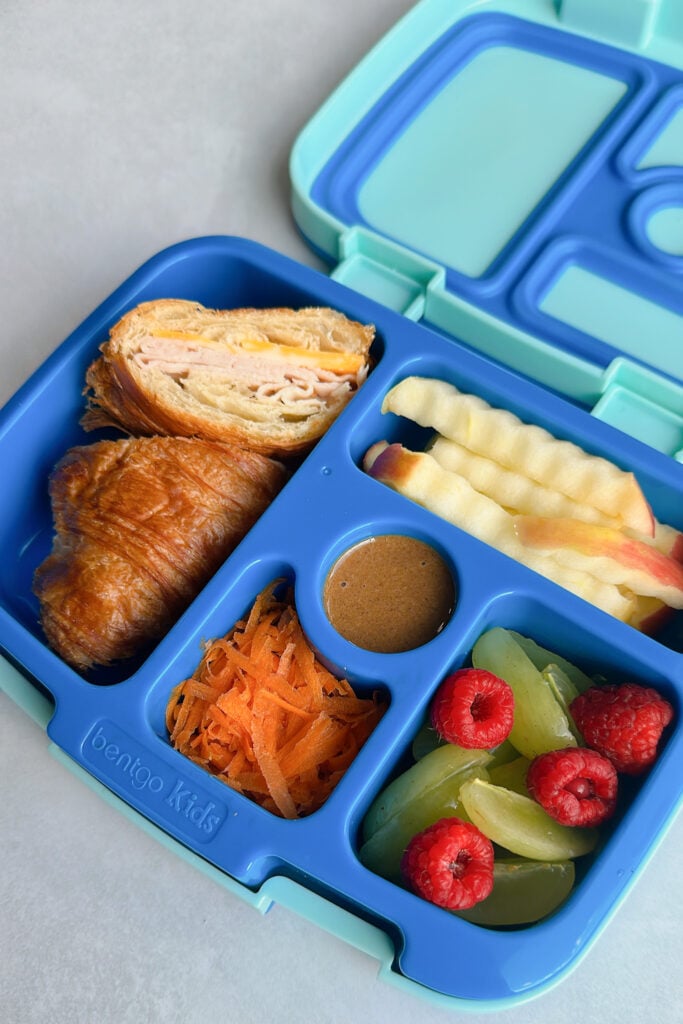 Turkey and cheese croissant served with apple's granola butter, grated carrots, grapes, and raspberries.