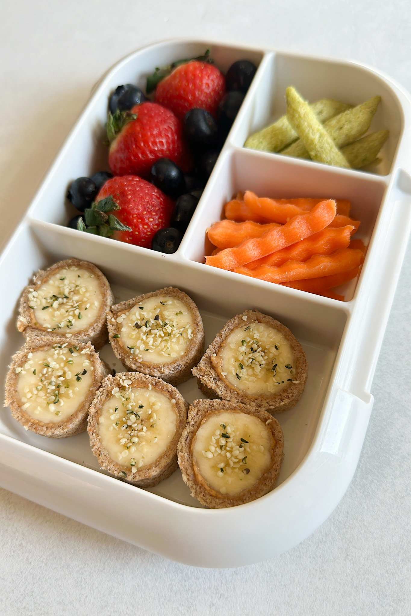 Kids lunchbox with banana sushi rolls, berries, carrots, and pea crisps.
