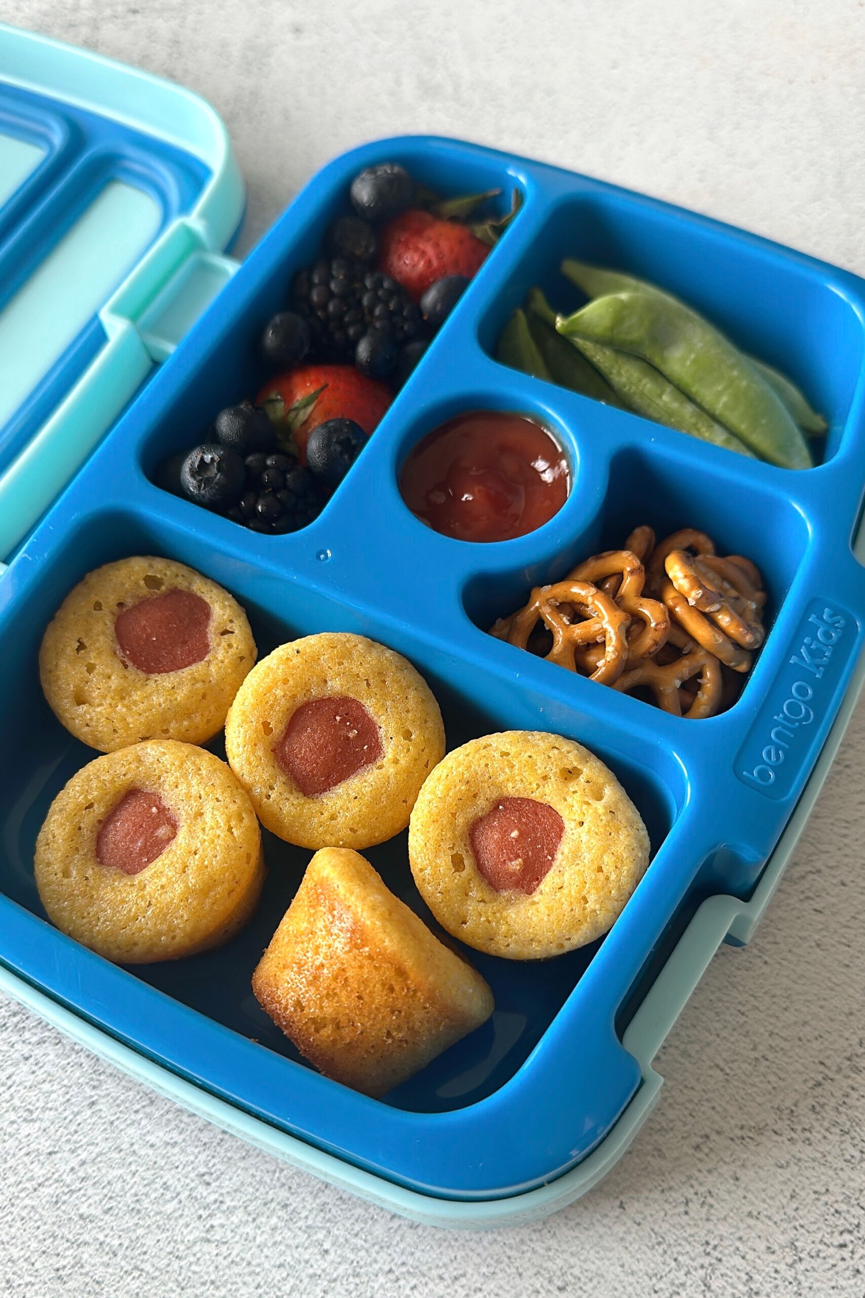 Corn dog bites served in a lunchbox with berries, pretzels, snap peas, and ketchup.